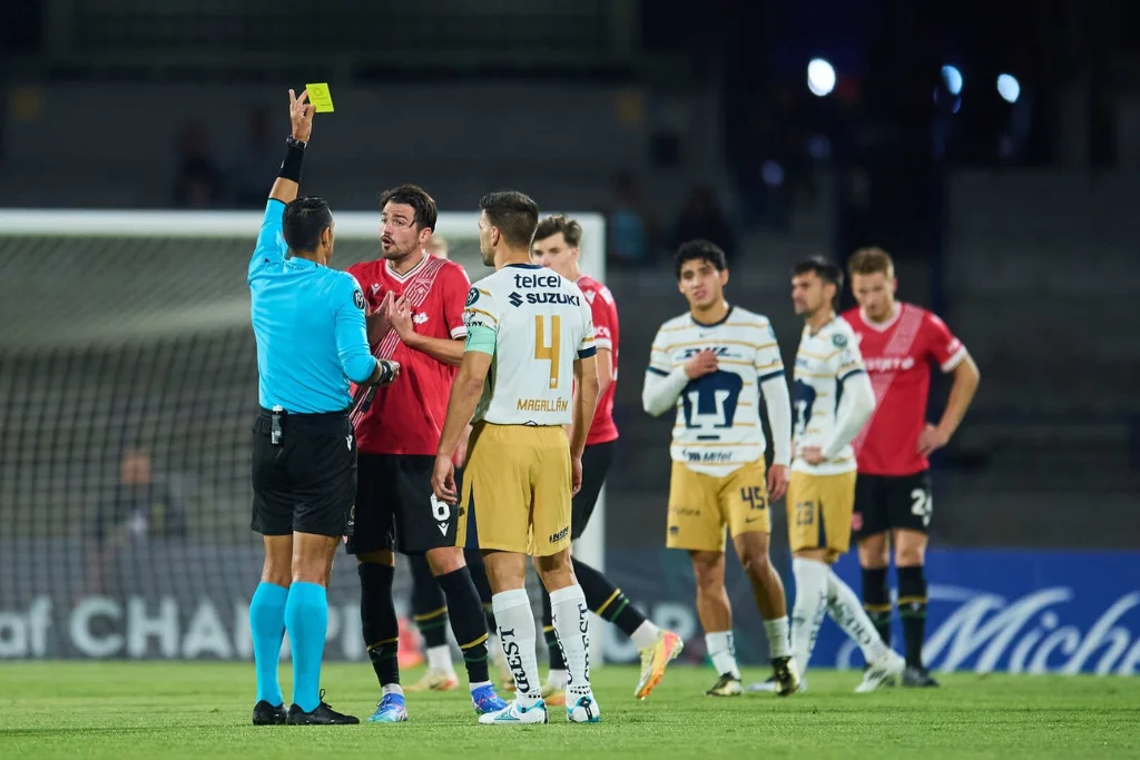 El árbitro Juan Calderón muestra tarjeta amarilla a un jugador del Cavalry, mientras Lisandro Magallán, observa la escena junto a sus compañeros en el fondo | Qué Onda