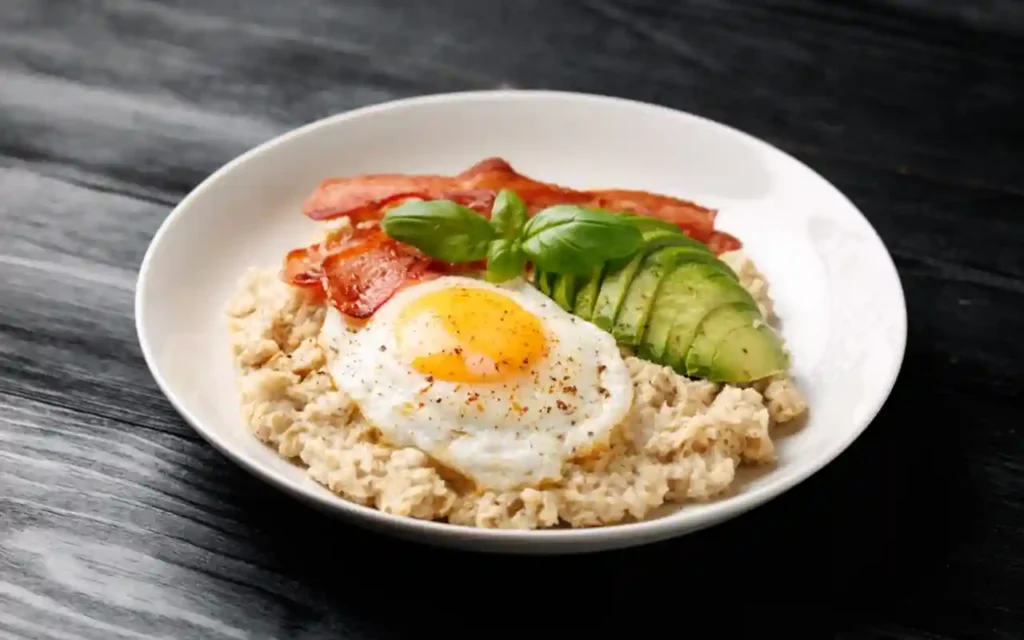 Avena preparada en versión salada, servida con un huevo frito, tiras de tocino crujiente y rodajas de aguacate fresco | Qué Onda