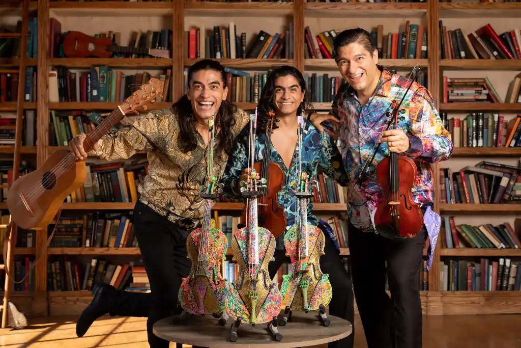 Villalobos Brothers posando de pie en una biblioteca con violines decorados con arte y guitarras acústicas | Qué Onda